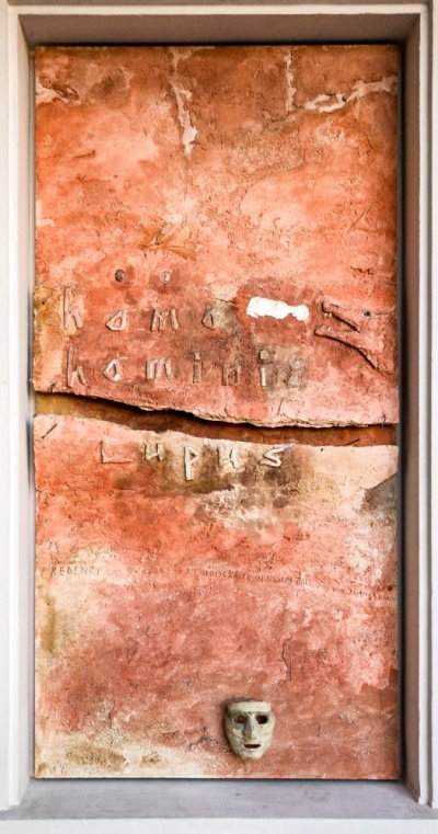 Bildtafel mit rötlichem Grund, einer Maske rechts unten und der als Relief ausgeführten Inschrift homo homini lupus